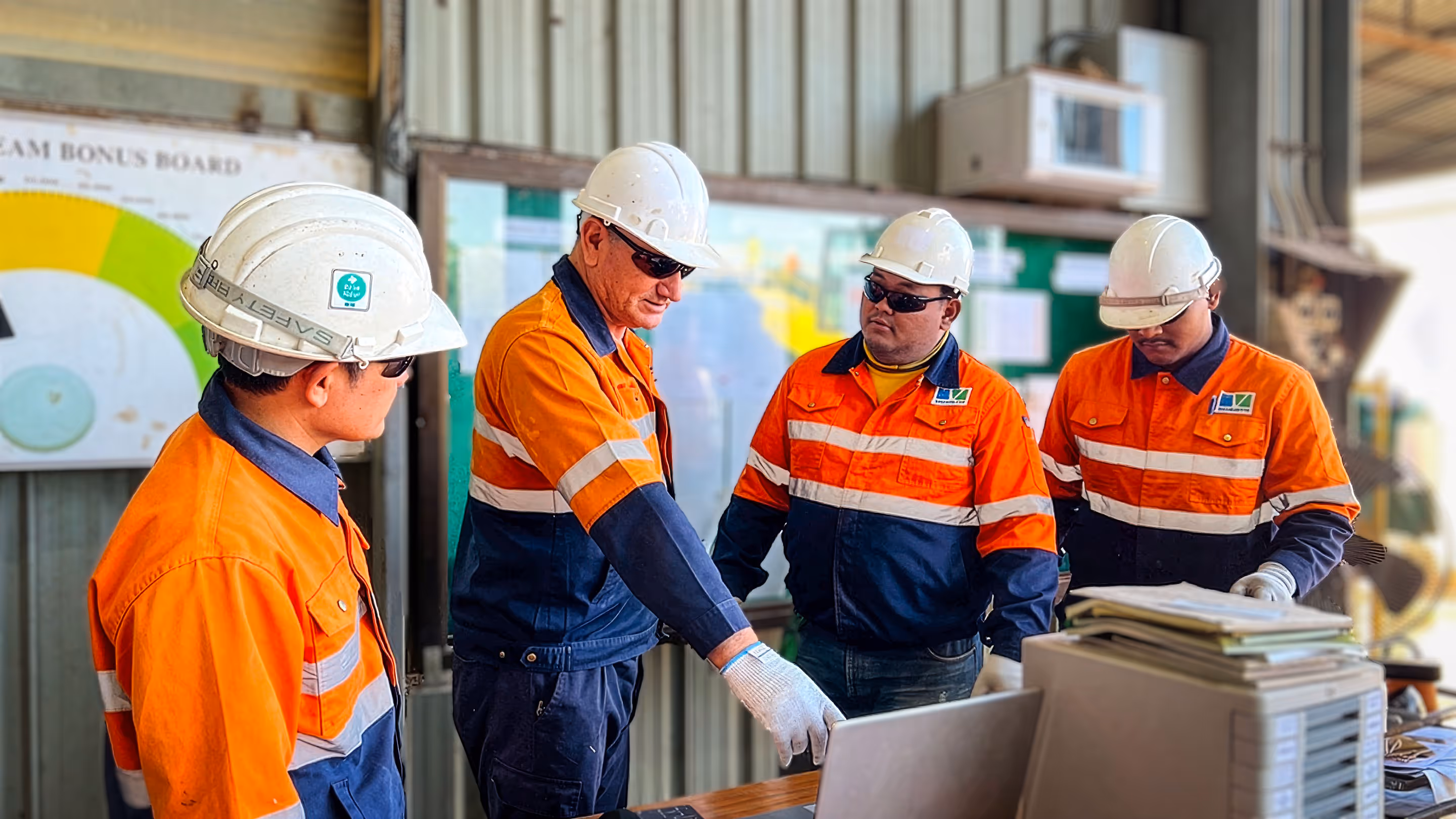 Four construction workers wearing orange and navy safety uniforms and white helmets gathered around a table, looking at a laptop and documents.