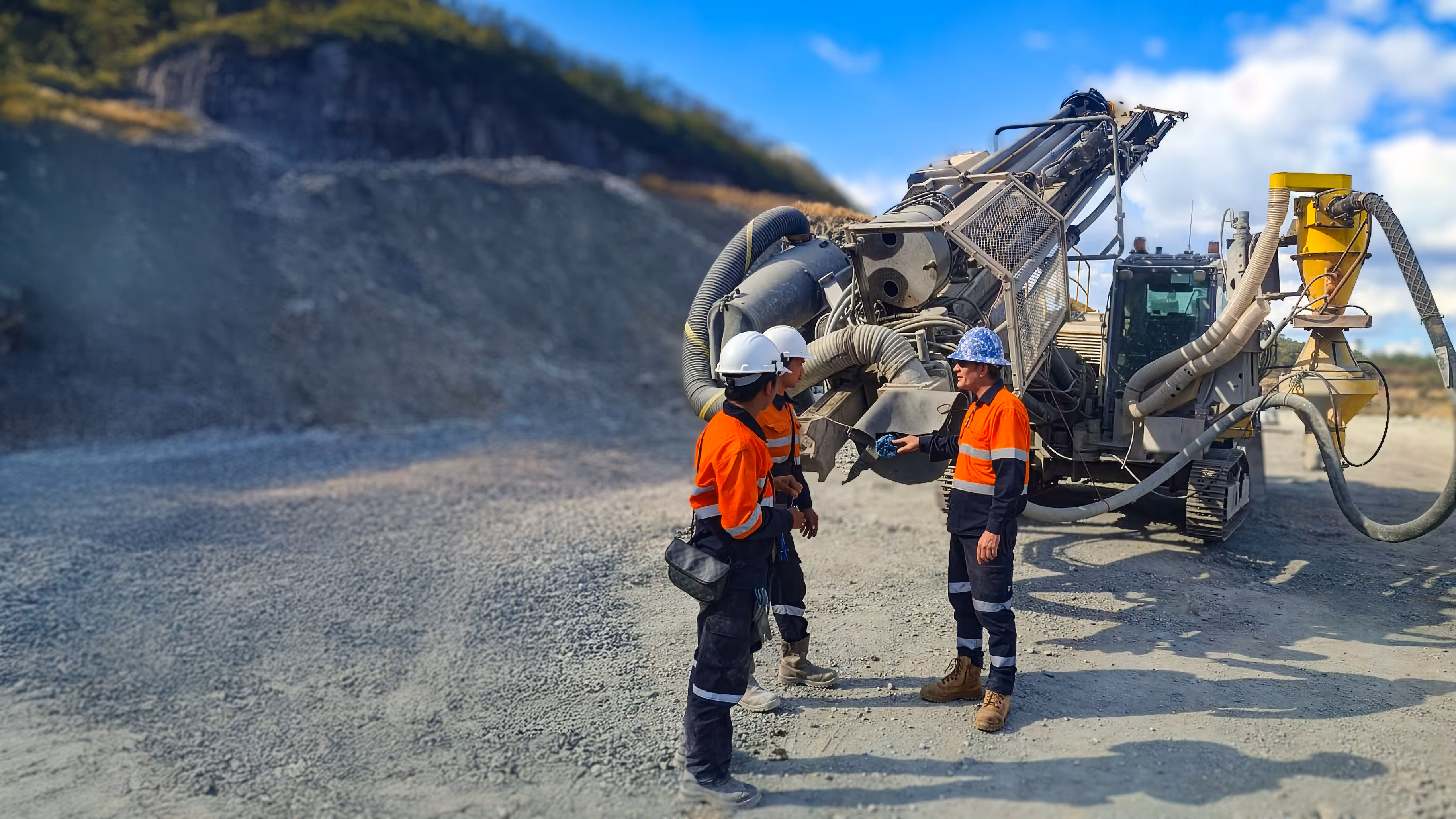 Three construction workers in orange safety jackets and helmets standing in front of large industrial machinery at a gravel or quarry site.