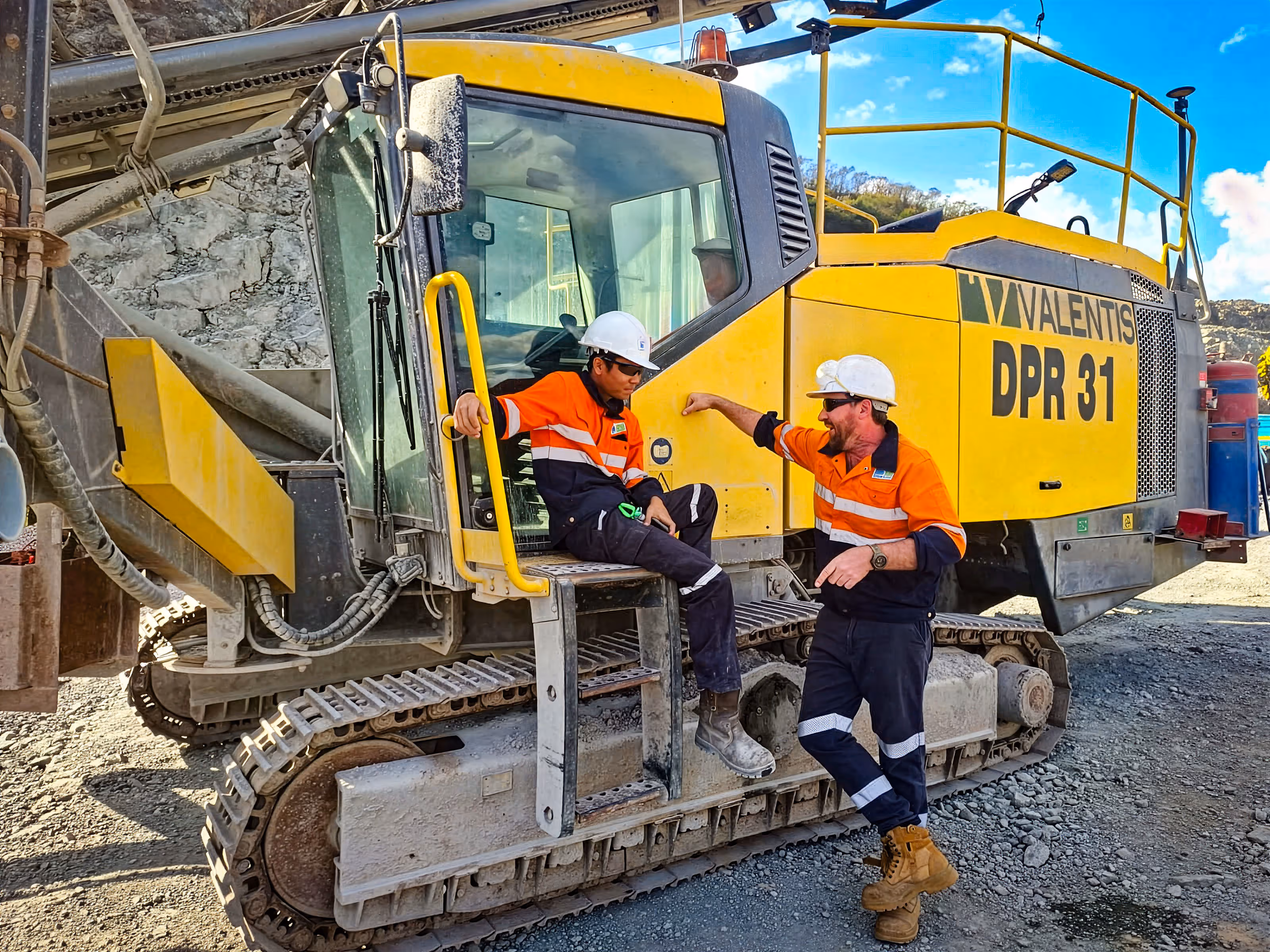 Two construction workers wearing orange and navy safety uniforms and white helmets talking beside and sitting on a large yellow tracked vehicle labeled VALENTIS DPR 31 at a gravel site.
