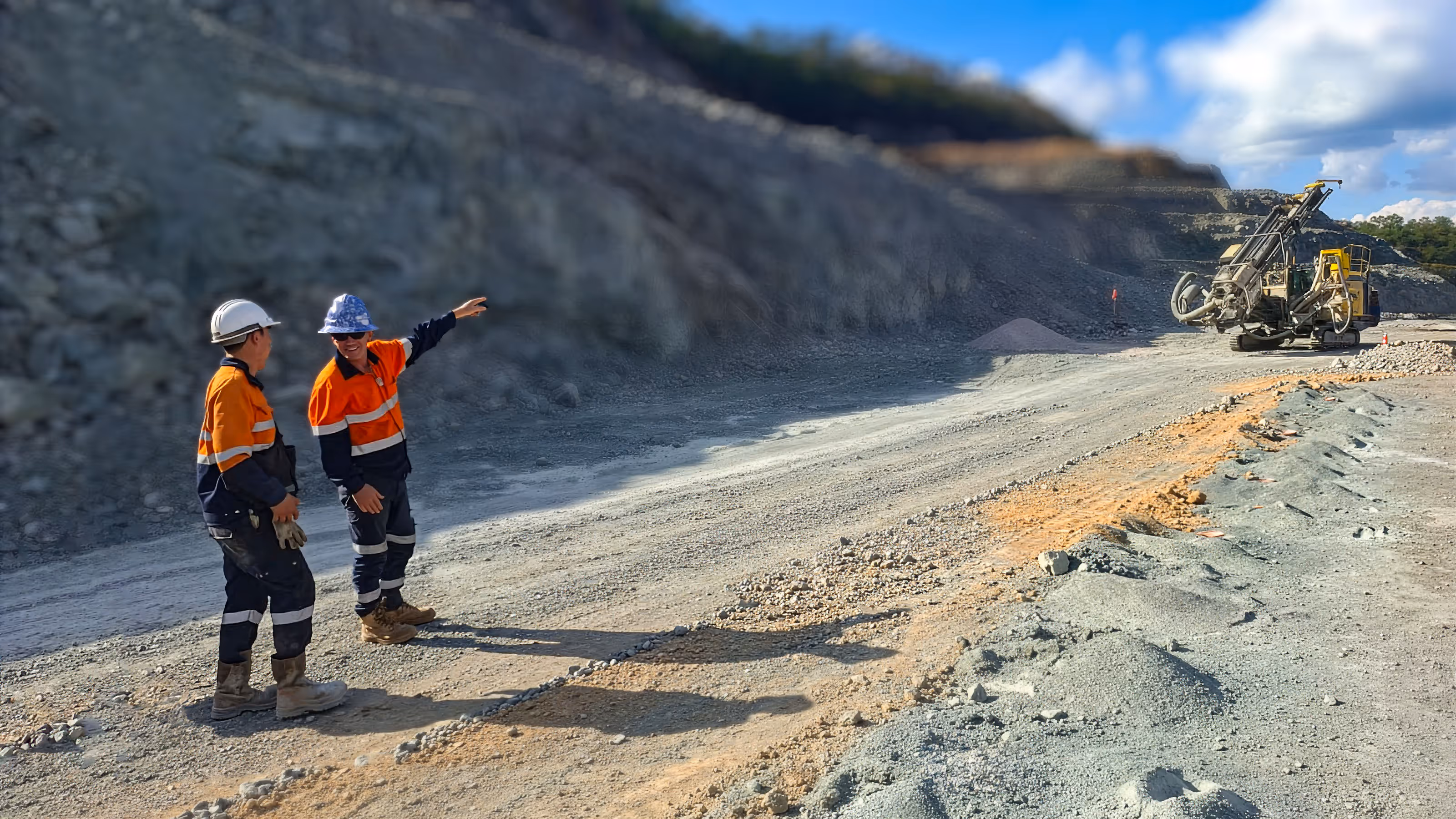 Two construction workers in orange safety gear and helmets standing on a gravel road in a quarry, one pointing toward drilling machinery in the distance.