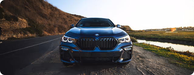 Front view of a blue BMW car parked on a dirt road beside a grassy field and calm water at sunset.