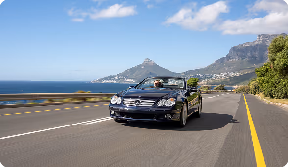 Black Mercedes convertible driving on coastal highway with mountains and ocean in the background on a sunny day.