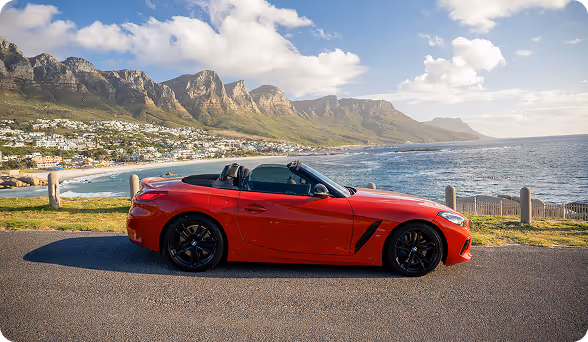 Red convertible sports car parked on a coastal road with mountains and ocean in the background.