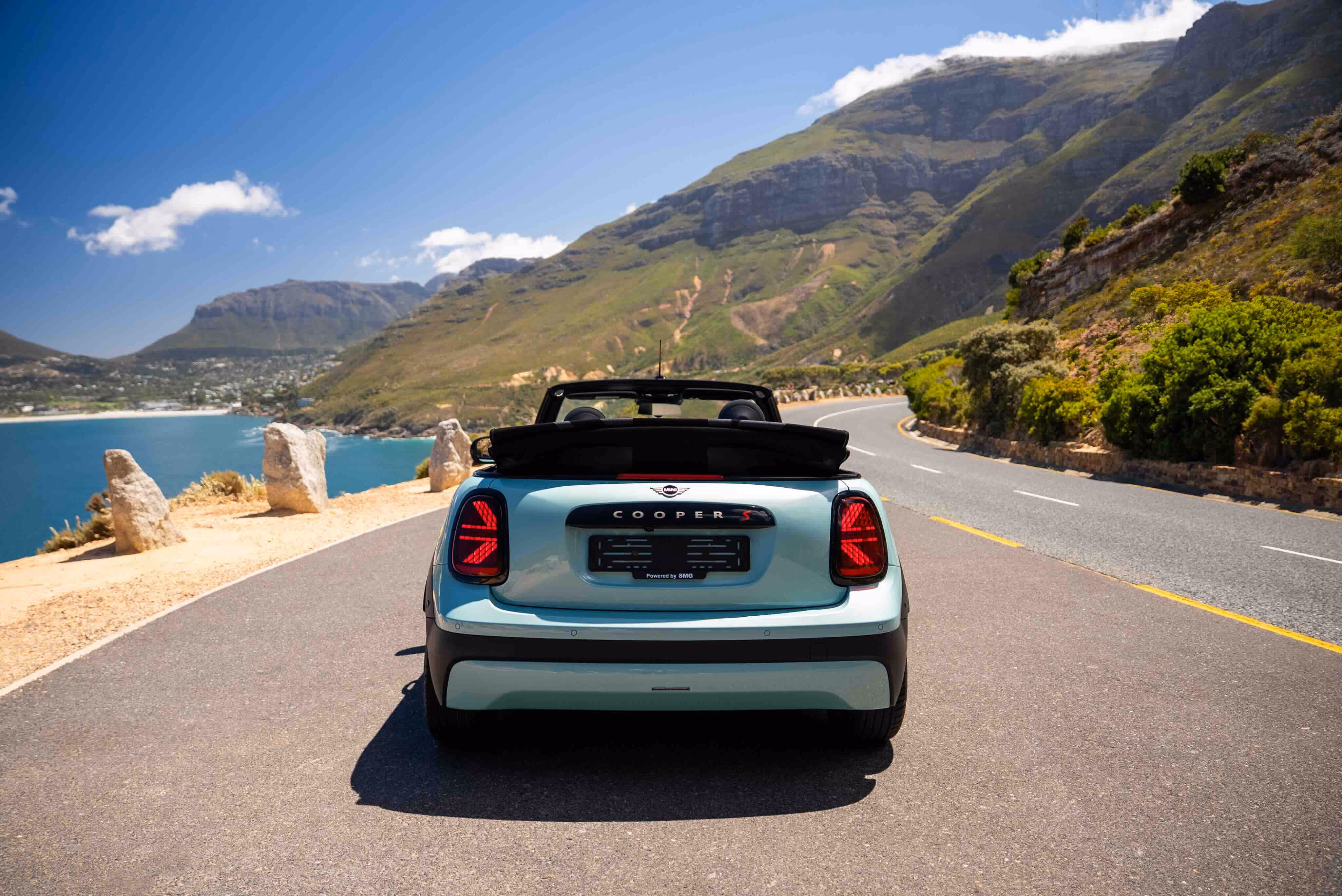 Rear view of a light blue Mini Cooper S convertible parked on a coastal road with mountains in the background and the ocean to the left under a blue sky with some clouds.