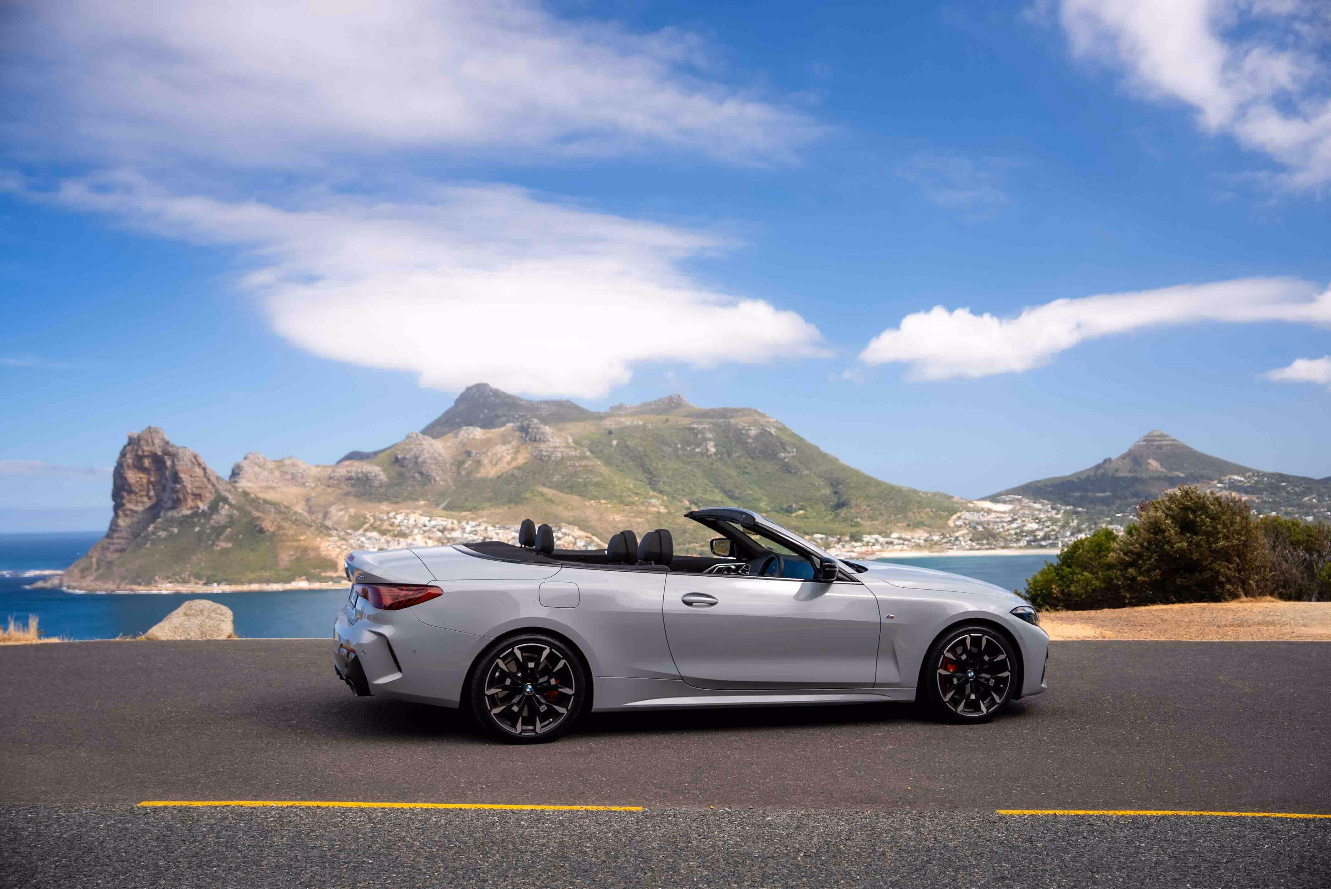Silver convertible sports car parked on a road with a scenic backdrop of green mountains, blue sky, and ocean coastline.