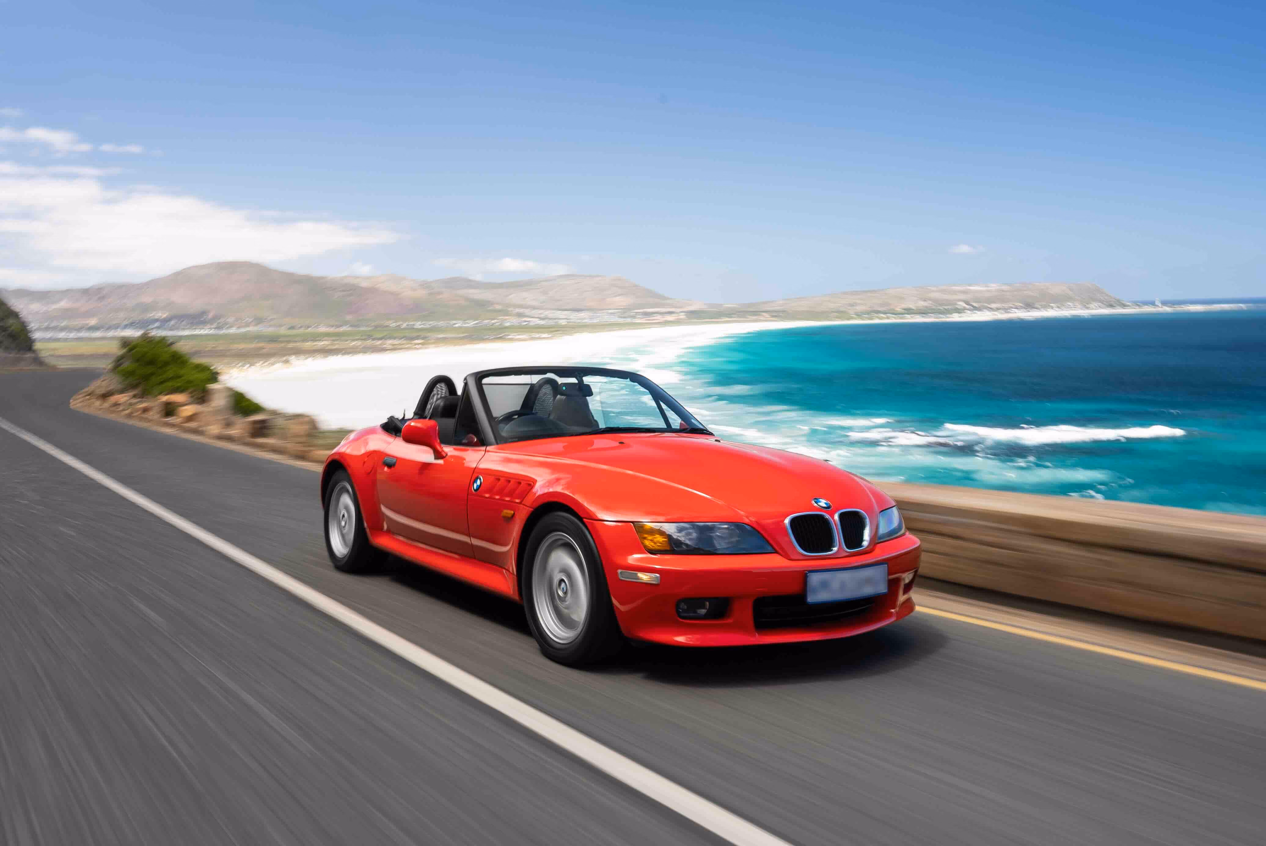 Red BMW convertible sports car driving on a coastal road with a scenic view of turquoise ocean, white sandy beach, and distant hills under a blue sky.