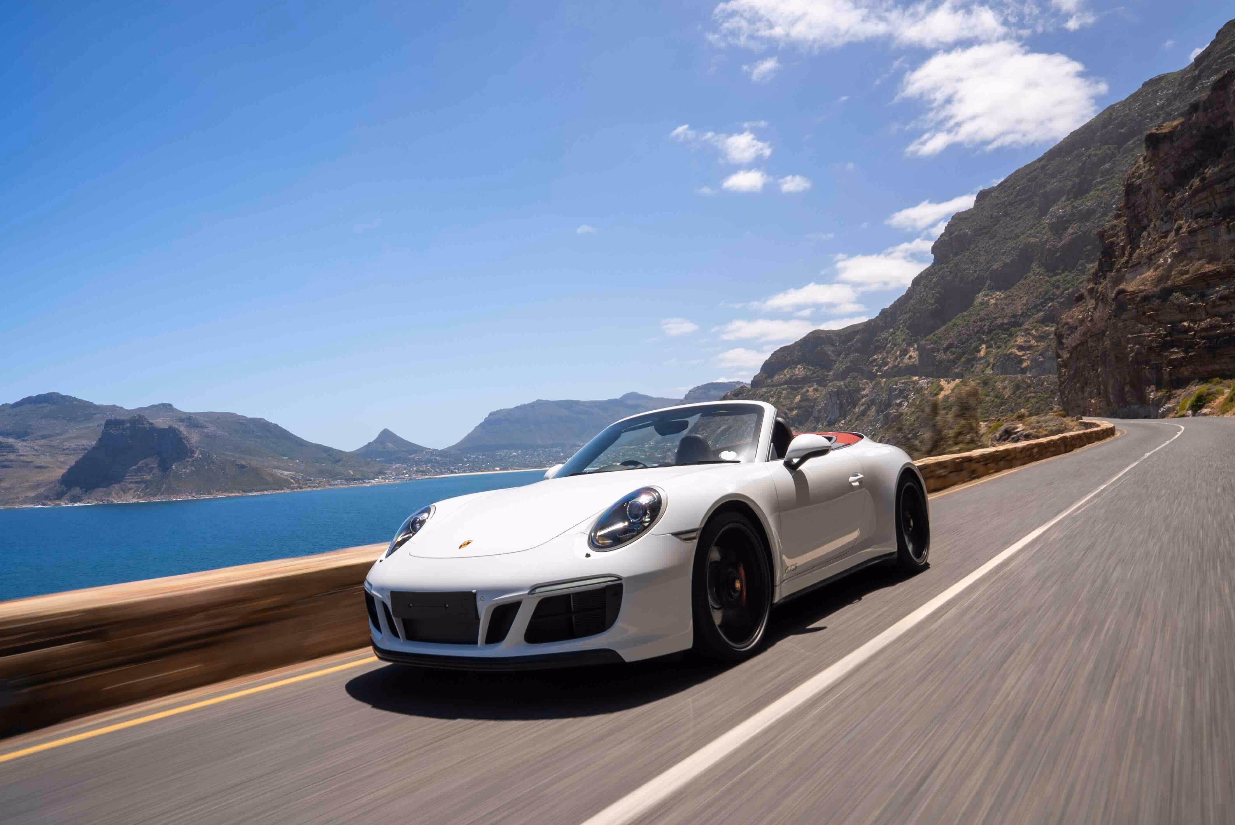 White Porsche convertible sports car driving on a coastal mountain road with blue ocean and rugged mountains in the background under a partly cloudy sky.