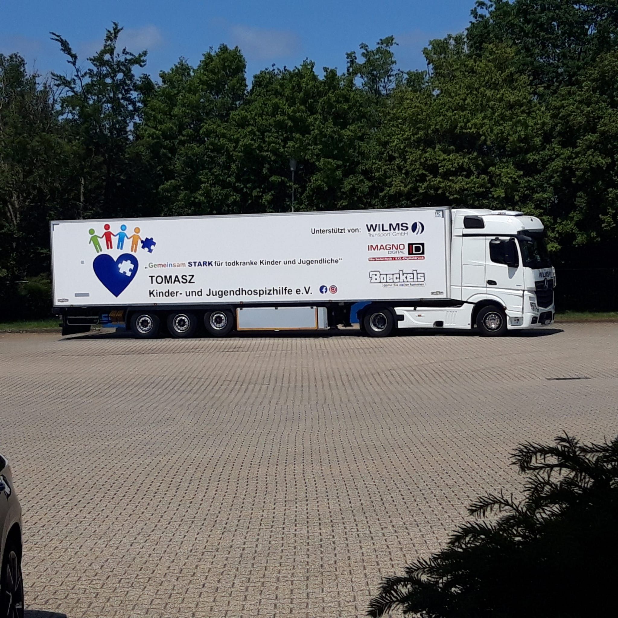 Weißer Lastwagen mit Beschriftung für TOMASZ Kinder- und Jugendhospizhilfe e.V. und buntem Herzlogo mit Puzzleteil, im Hintergrund Bäume und blauer Himmel.