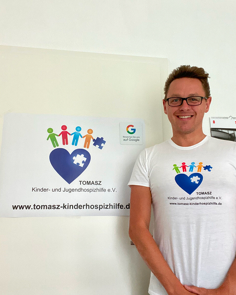 Ein Mann mit Brille trägt ein weißes T-Shirt mit dem Logo von Tomasz Kinder- und Jugendhospizhilfe e.V., neben ihm ein Plakat mit demselben Logo an der Wand.