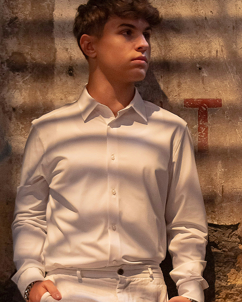 Junger Mann in weißem Hemd steht vor einer rauen Wand mit Schattenstreifen und blickt nach rechts.