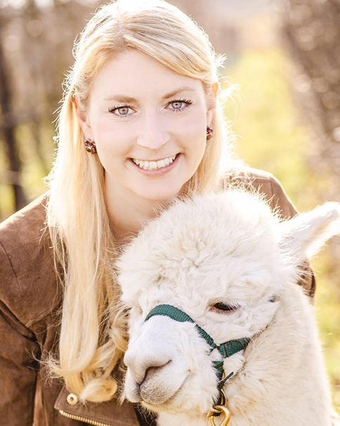 Lächelnde blonde Frau mit langem Haar neben einem weißen Alpaka mit grünem Halfter.