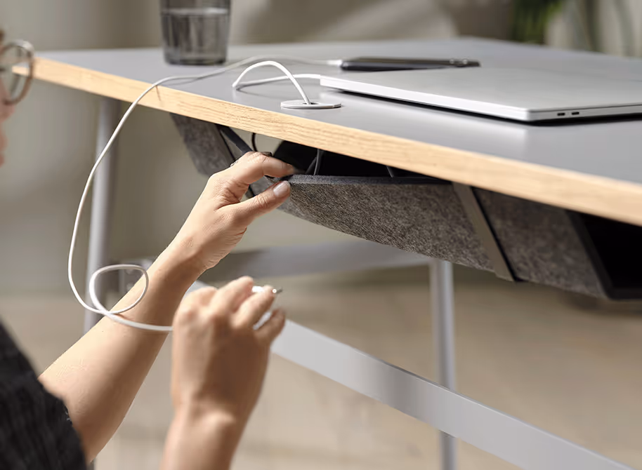 Een persoon die een grijze kabelgoot onder een bureau vastmaakt terwijl een laptop en een glas water op het blad staan.