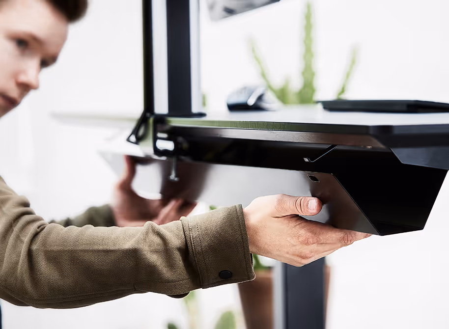 Een persoon installeert of bevestigt een zwart metalen onderdeel aan een meubelstuk of apparaat in een heldere ruimte met planten op de achtergrond.