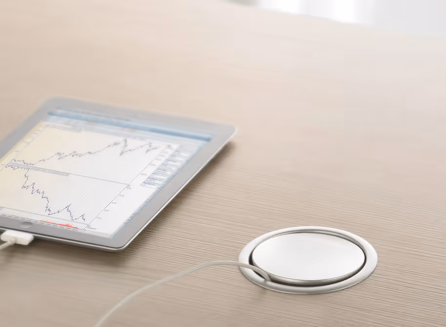 Tablet op een houten bureau met grafiek op het scherm en een verbonden witte oplaadkabel in een metalen inbouw stekkerdoos.
