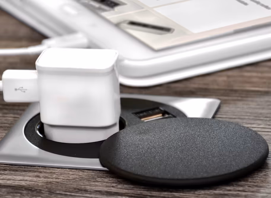Witte USB-lader verbonden met een ingebouwd stopcontact op een houten bureau, naast een zwart rond object en een deel van een laptop.