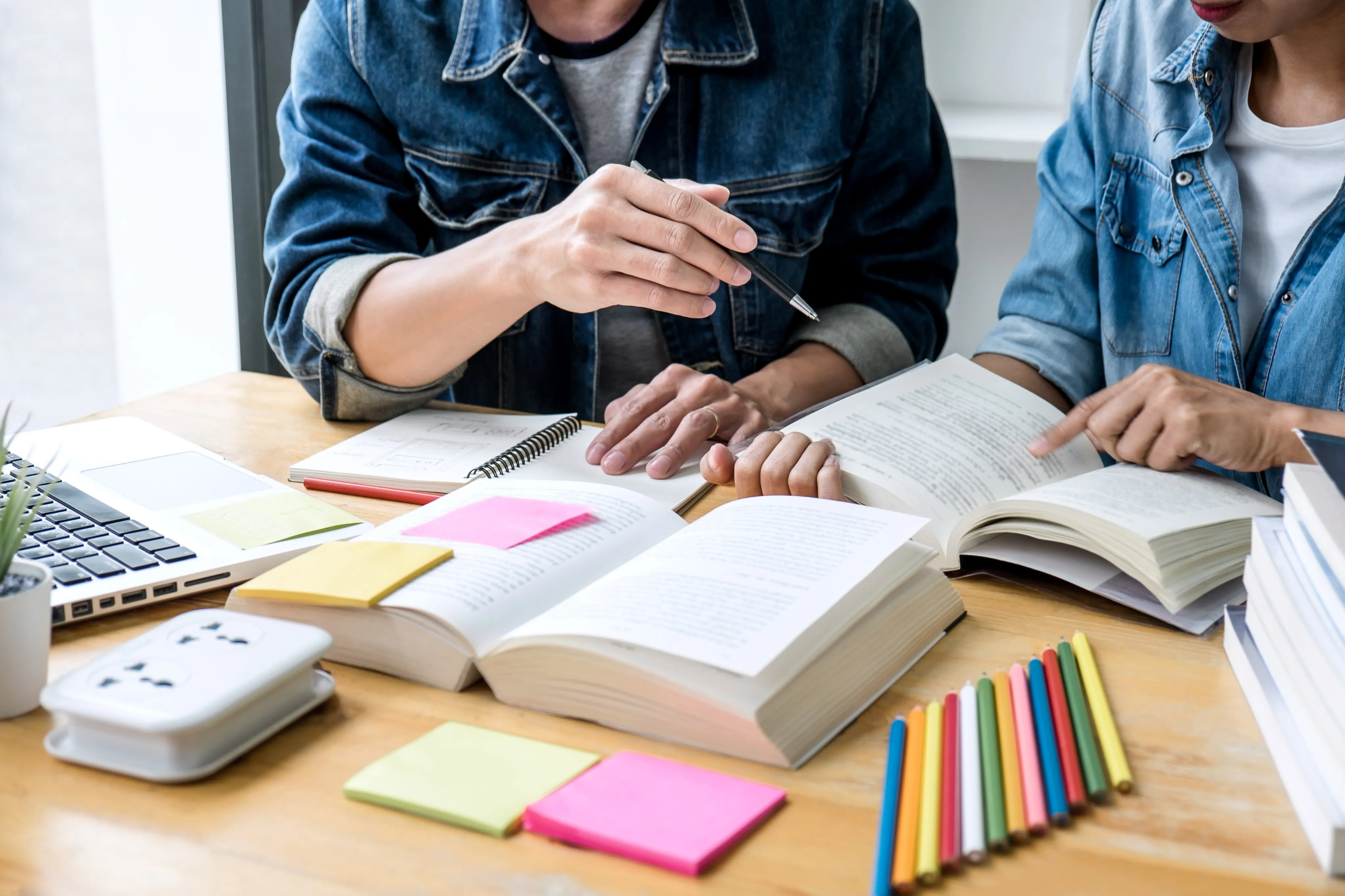 Two people in denim jackets studying together with open books, colored pencils, sticky notes, and a laptop on a wooden table.