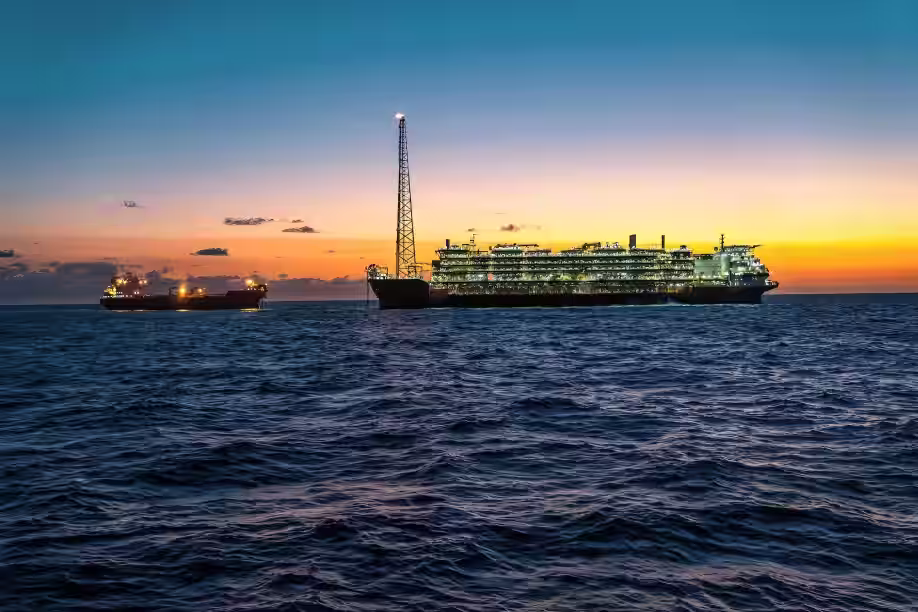 Large illuminated ship with a tall structure and a smaller boat on calm sea during sunset.