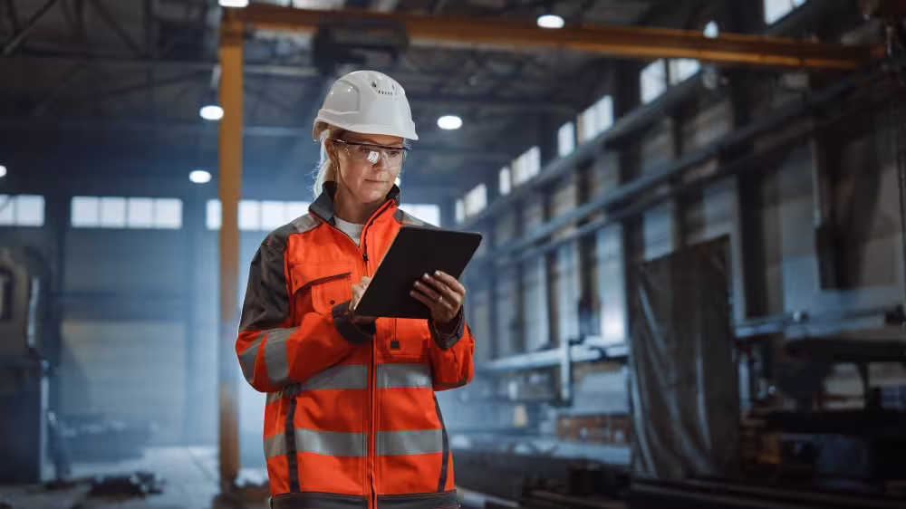 Project assurance professional in safety helmet and orange reflective jacket using a tablet in an industrial warehouse.
