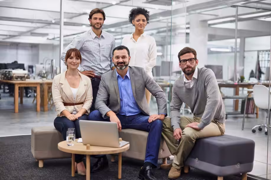 Graham Sutherland with his team of diverse project assurance professionals posing and smiling in a modern office with glass walls and wooden desks.