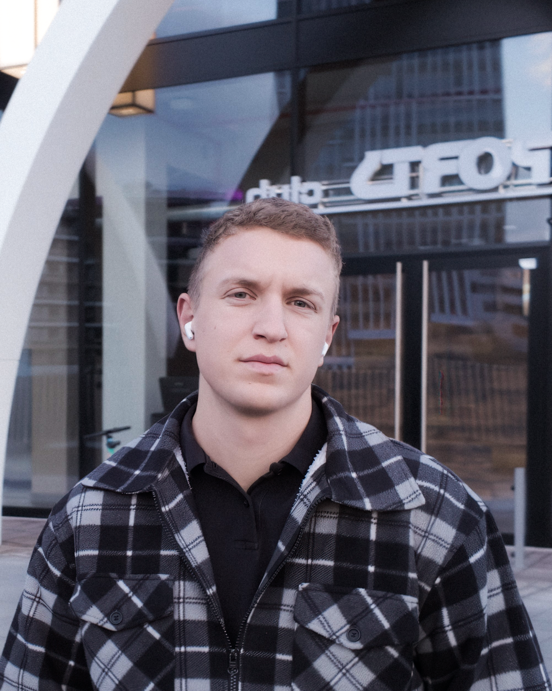 Porträt eines jungen Mannes mit kurzen blonden Haaren und kariertem Mantel, der vor einem Gebäude mit Glasfassade steht.