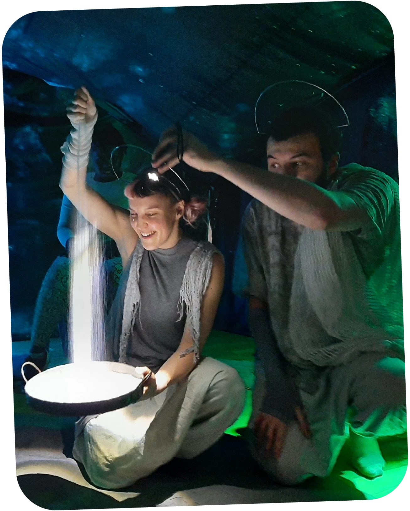 Two people kneeling indoors, one smiling and pouring a bright stream of light or fine sand from a pan.