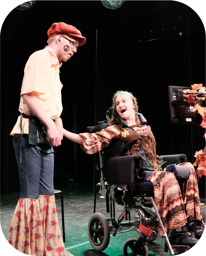 A young woman in a wheelchair holding hands and smiling at a young man dressed in colorful, retro-style clothing on stage.