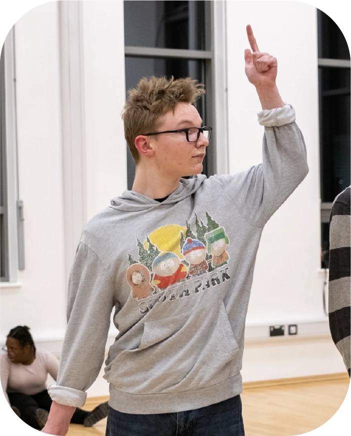 Young man wearing glasses and a grey hoodie raising his right arm with index finger pointed up indoors.