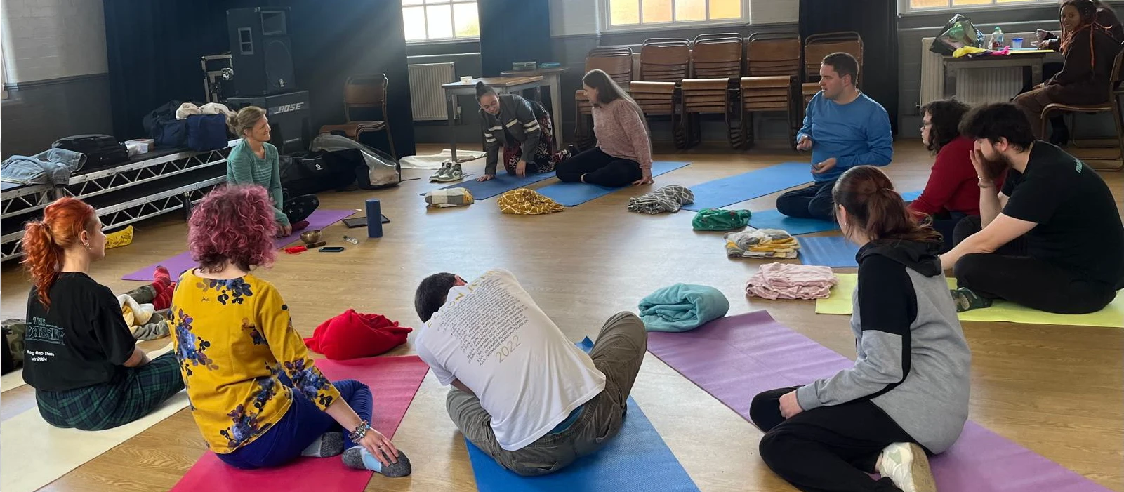 Group of young adults seated on yoga mats in a spacious room, engaging in a yoga class
