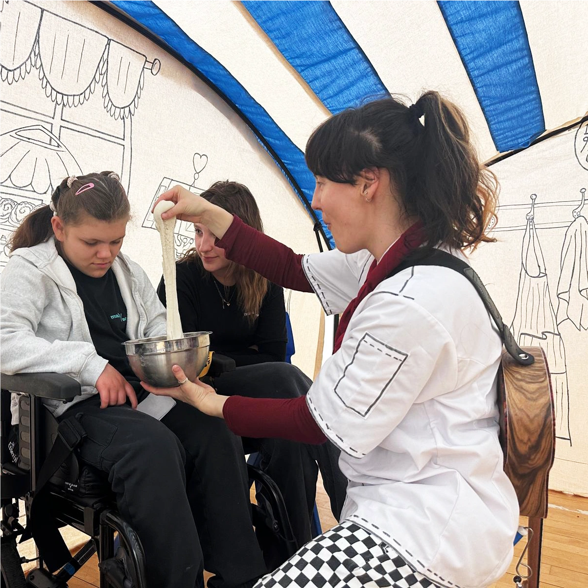 Woman demonstrating stretchy dough with a metal bowl to a seated young person in a wheelchair, inside a tent with drawn decorations of a bakery setting.
