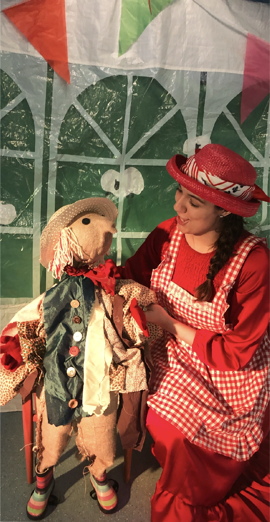 Woman in a red dress and gingham apron with a red hat interacting with a colorful puppet scarecrow dressed in patchwork clothes.