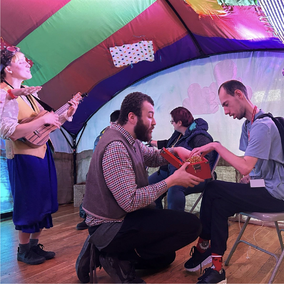 Woman dresses as a pirate playing a ukelele under a colorful tent while a man kneels presenting a treasure chest with golden paper to a seated young man.