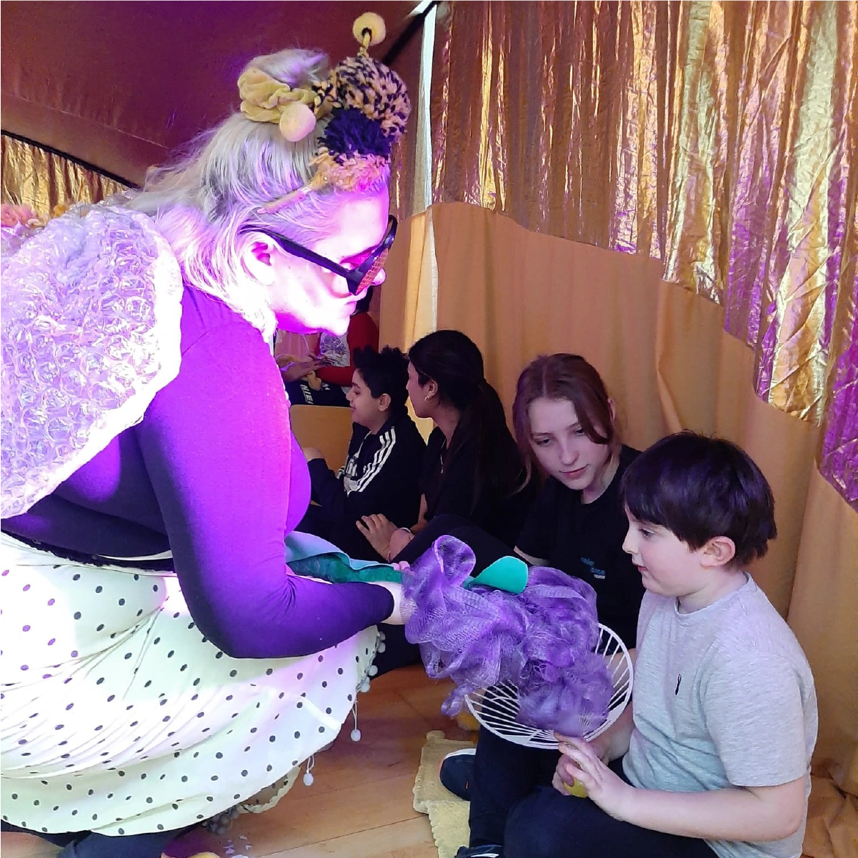 Woman dressed in bee costume with wings and an antenna headband interacts with two children sitting and holding a lavender flower prop.