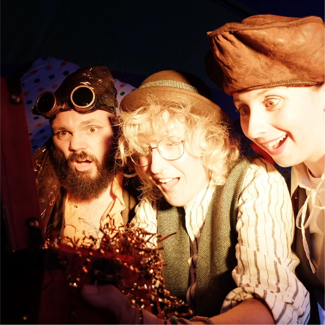 Three people in pirate costumes looking excitedly at a treasure chest filled with shiny objects.