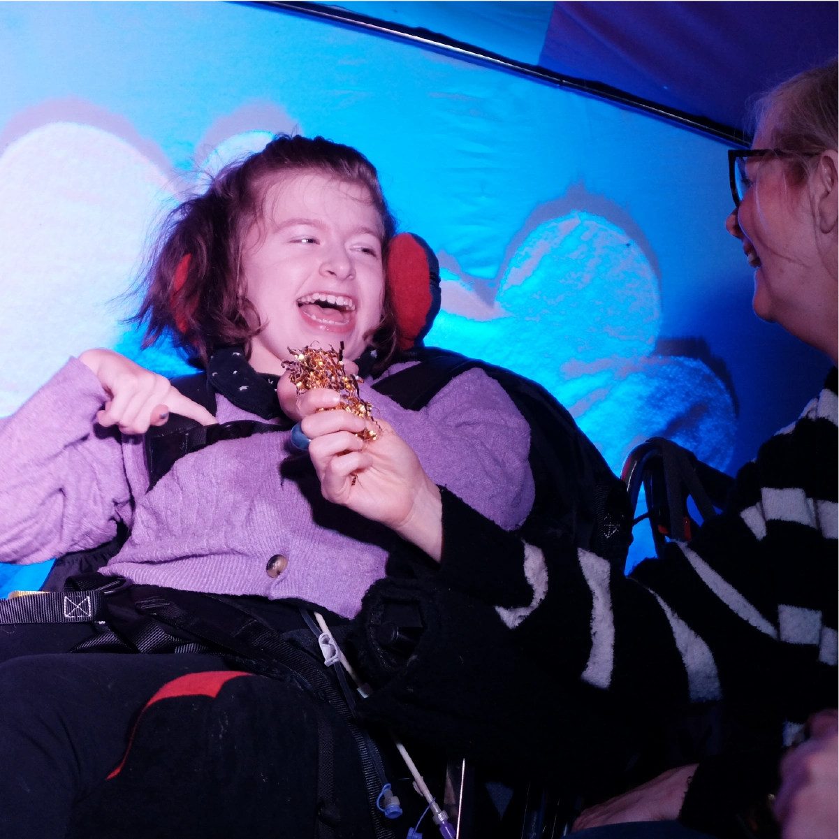 Smiling young girl wearing a purple sweater, seated in a wheelchair, laughing and holding decorated confetti with a woman smiling beside her.