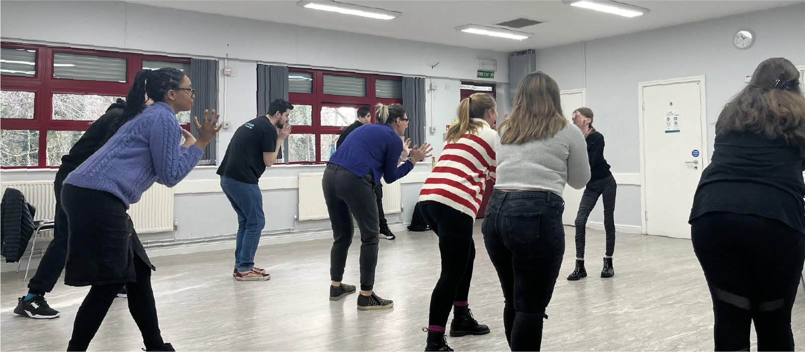 Group of young people standing in a semi-circle in a bright room, leaning forward with hands near their faces, engaged in a group workshop.