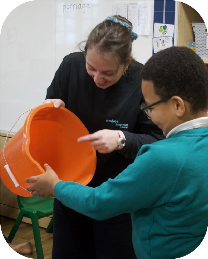 A woman and a boy smiling as they hold an orange bucket together in a classroom setting.