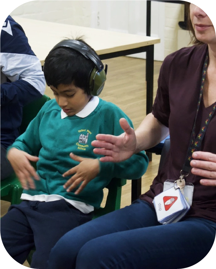 A young student wearing ear defenders and a green school sweatshirt sits next to an adult who is gesturing with their hands during a warm up game.