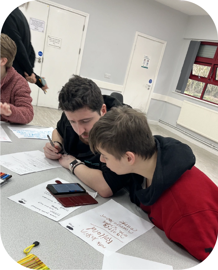 A staff member assists a young person, leaning over a table, discussing notes on paper and a smartphone.