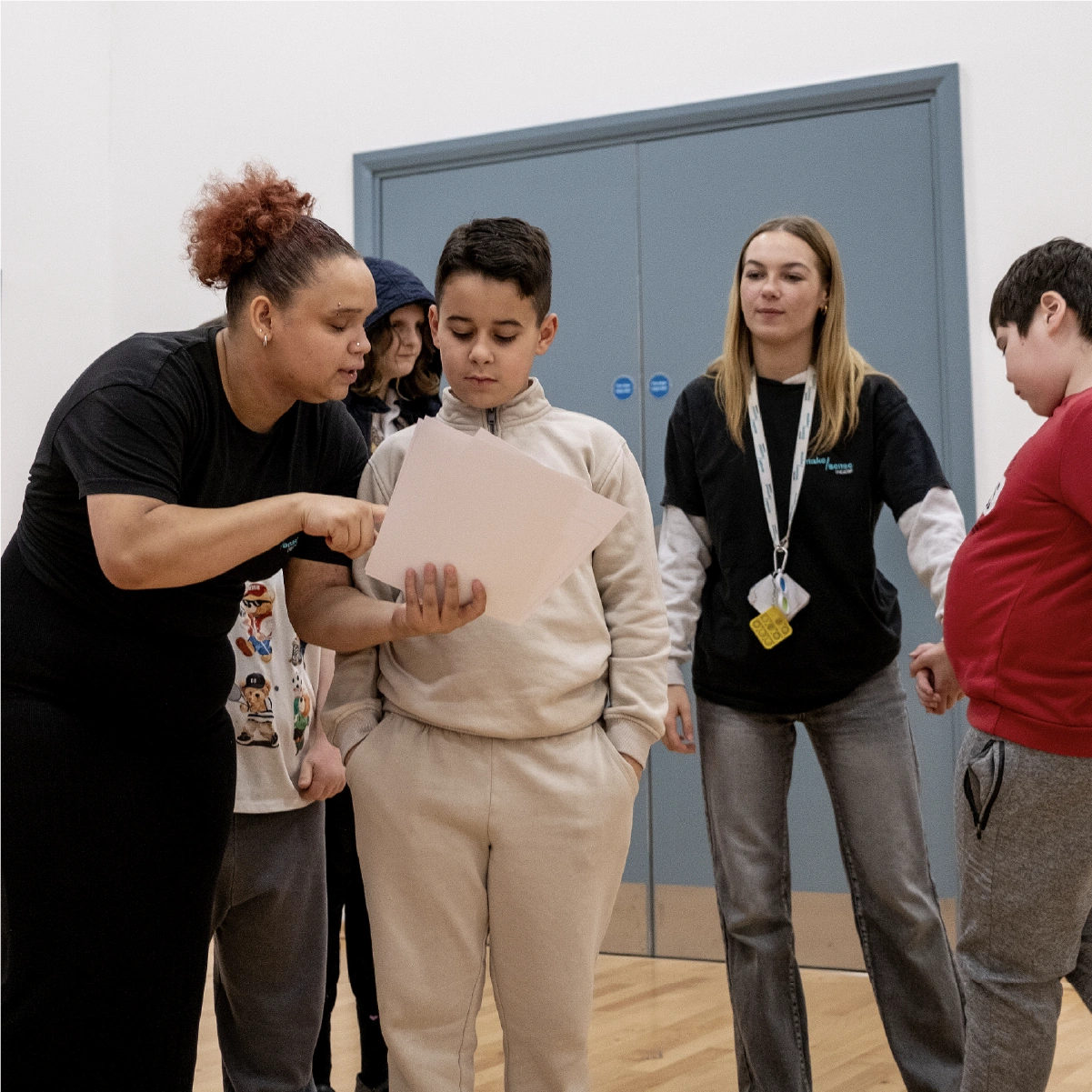 A woman in a black shirt shows papers to a boy in beige clothes while two other children and a woman stand nearby indoors
