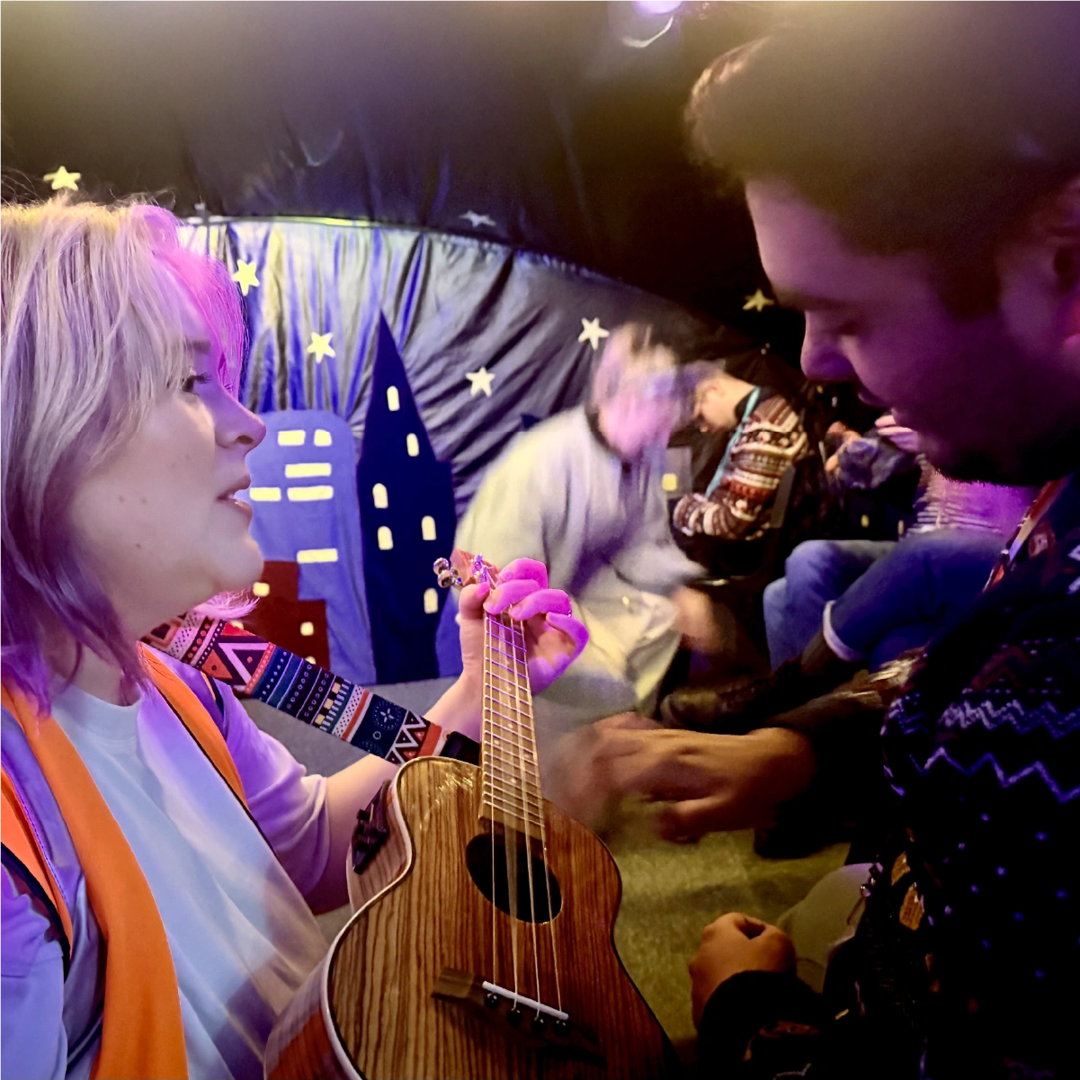 A woman playing a wooden ukulele while a young person watches closely in a tent with a night sky-themed backdrop and other people seated in the background.