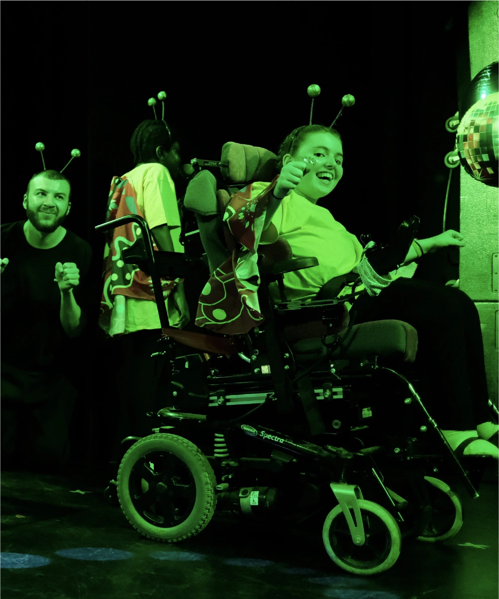 Three people, including a girl in a wheelchair wearing a yellow shirt and butterfly wings, dressed with antenna headbands, smiling and giving thumbs up under green lighting.