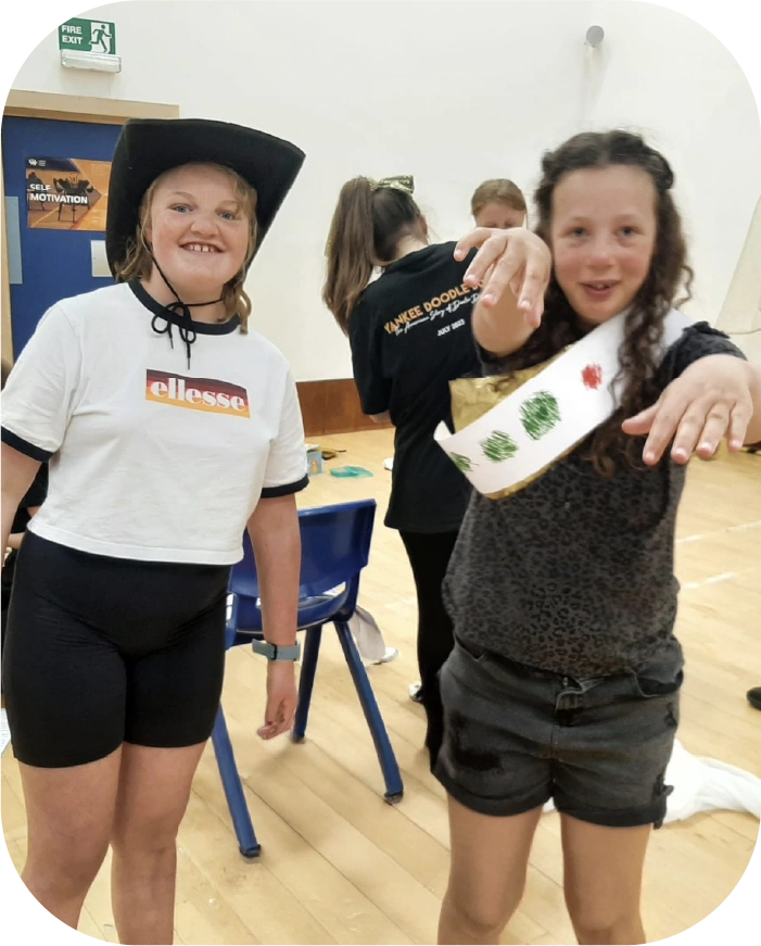 Two young girls indoors, one wearing a black hat and white t-shirt, the other with curly hair wearing a sash with green and red decorations posing with ams stretchd in front of her