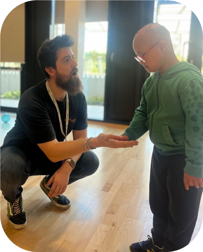 Man with a beard crouching indoors, holding out his hand to a child in a green hoodie and glasses.