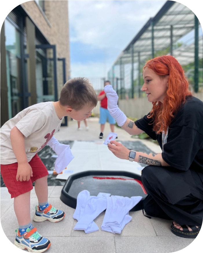 A woman with red hair wearing black clothes demonstrates sock puppets to a young boy in a white shirt and red shorts outdoors.