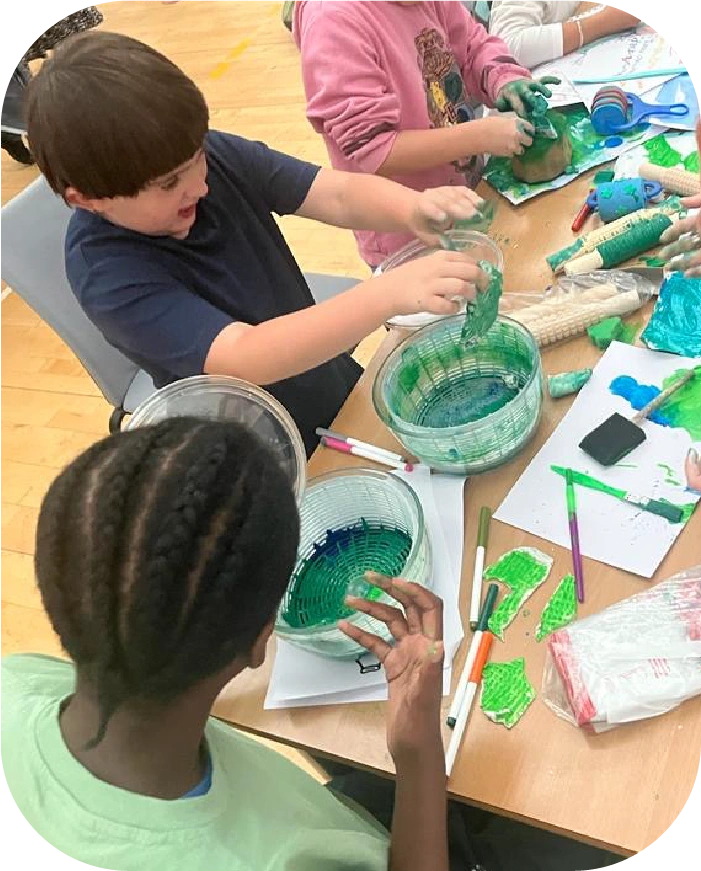 Children sitting around a table engaging in a green paint craft activity with bowls of green paint and art supplies scattered on the table.