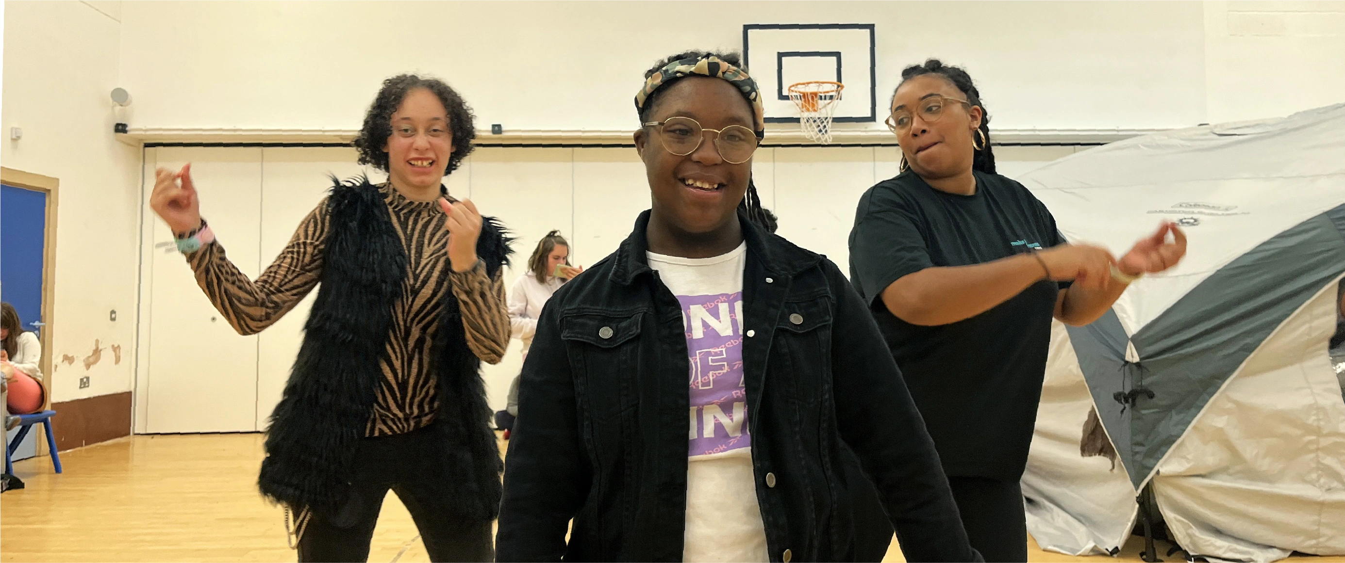 Three smiling young people dancing indoors with a tent in the background.