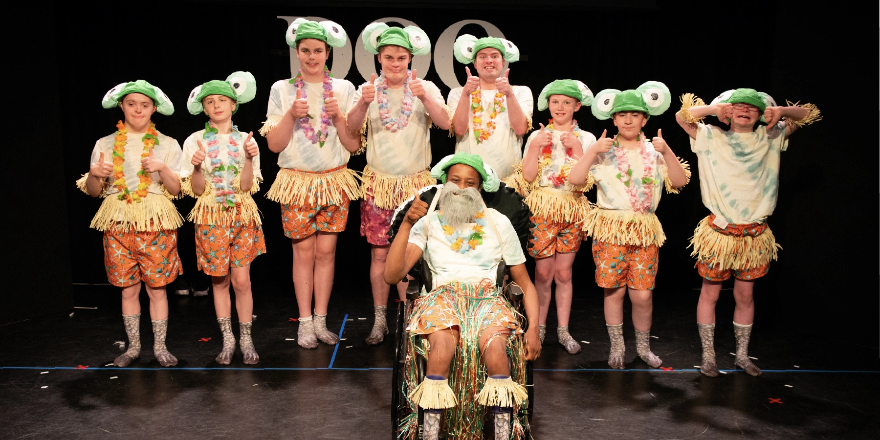 Group of young people wearing turtle hats, floral leis, grass skirts, and starfish-patterned shorts posing on stage with thumbs up, one young person in a wheelchair dressed similarly.