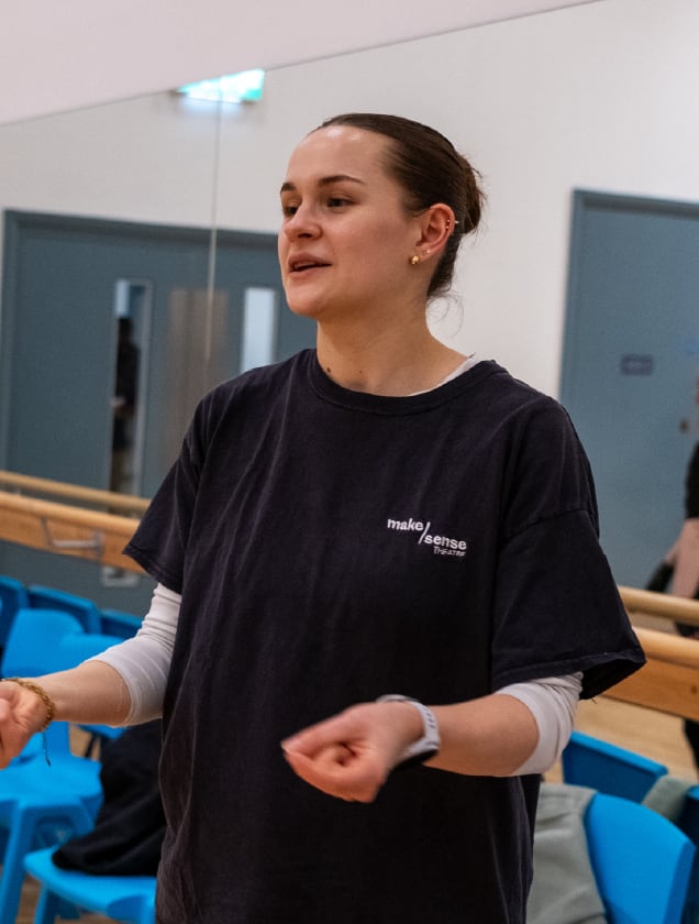 Young woman wearing a black 'make sense theatre' T-shirt speaking in a room with blue chairs and mirrors.