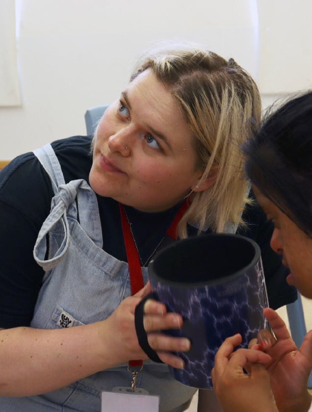A woman with blonde hair and a nose piercing holds a drum while looking up, sitting beside another person.