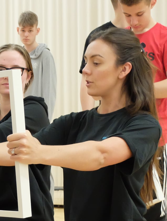 Woman holding a white rectangular picture frame horizontally in a room with other people in the background.